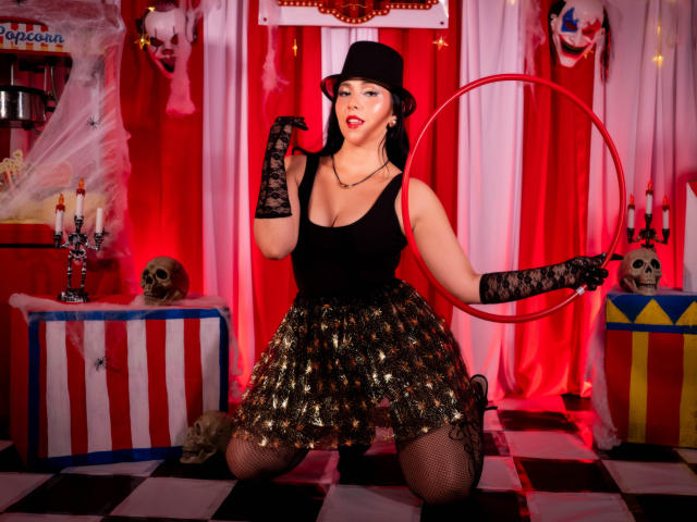 Katty Rivera mit schwarzem Hut, Spitzenhandschuhen, goldfarbenem paillettenbesetztem Rock, schwarzem Tanktop und Fischernetzstrümpfen, kniend, einen Reifen haltend, mit rotem Lippenstift, die ihre großen Brüste zeigt, sexy, im Halloween-Stil..
