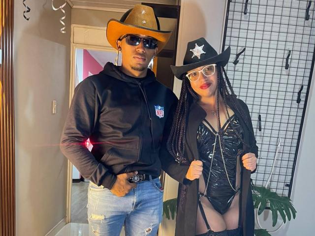 LilithAndAsmodeo, con un sombrero de vaquero y una sudadera negra con jeans azules, está de pie junto a LilithAndAsmodeo, que lleva un conjunto de cuero negro, un sombrero de vaquero y gafas, mostrando su vulva..