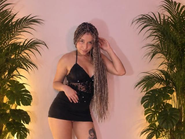 AtheneaClark with long braided hair, black halter mini dress, showing her tits, ass, and tattoo, standing between green plants, hand in hair, smiling, soft pink background.