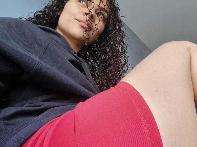MissSunnyShine, avec ses cheveux noirs bouclés, portant un sweat à capuche noir et un short rouge, montrant ses cuisses nues et légèrement poilues, dans une prise de vue en contre-plongée. Ses lèvres sont légèrement pincées..