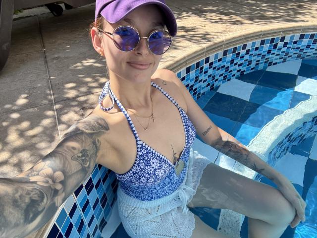 SarahRousen in a blue floral bikini top, white lace shorts, purple cap, round sunglasses, tattoos, sitting in a blue-tiled pool, smiling at the camera, with water reflections.