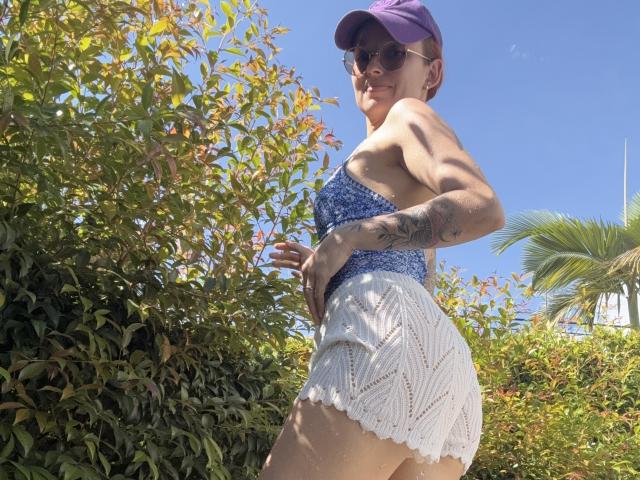 SarahRousen with short red hair, purple cap, sunglasses, blue patterned top, white lace shorts, tattoo on right arm, standing outdoors, looking back, clear blue sky, green bush.