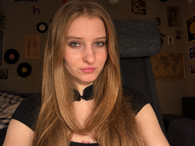 Ardelle Cooksey, met lang bruin haar, groene ogen en roze lippen, draagt een zwart shirt met een kraag en zit in een donkergrijze bureaustoel, met een lichte frons..