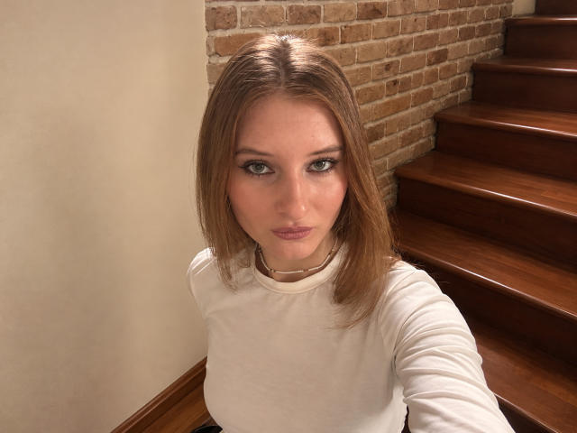 ArdelleCooksey with medium brown hair, green eyes, and white shirt, looking up at the camera, standing on wooden stairs, brick wall in background, necklace visible.