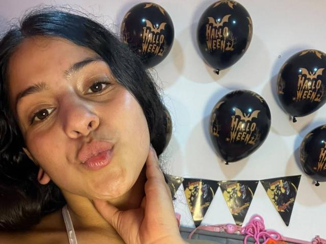 LonxEva with puffy lips, brown eyes, and long black hair, holding her neck, surrounded by black "Mandalore Weekend" balloons, wearing a pink bra, and pink ribbon.