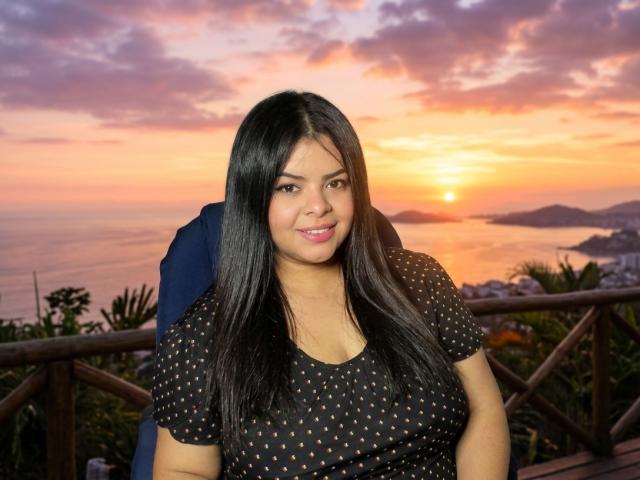 CandyValentine, con cabello largo y negro, vestida con una camisa negra de lunares, sentada al aire libre al atardecer, sonriendo, mostrando sus pechos y mirando a la cámara..