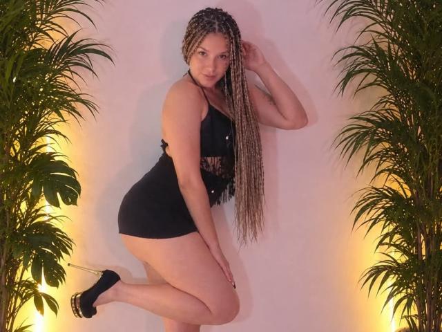 AtheneaClark with long blonde braids, wearing a black tank top and mini skirt, black high heels, standing on one leg, hands near head, green plants on sides.