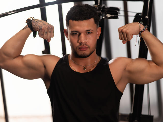 MasonCole, ein muskulöser Mann mit kurzen, schwarzen Haaren, der seine Arme anspannt, trägt ein schwarzes Tanktop, hat ein Tattoo auf der rechten Hand, ein Armband am linken Handgelenk, einen ernsten Gesichtsausdruck, sichtbare Bizeps, eine Halskette und hat braune Haut. Er befindet sich im Fitnessstudio..