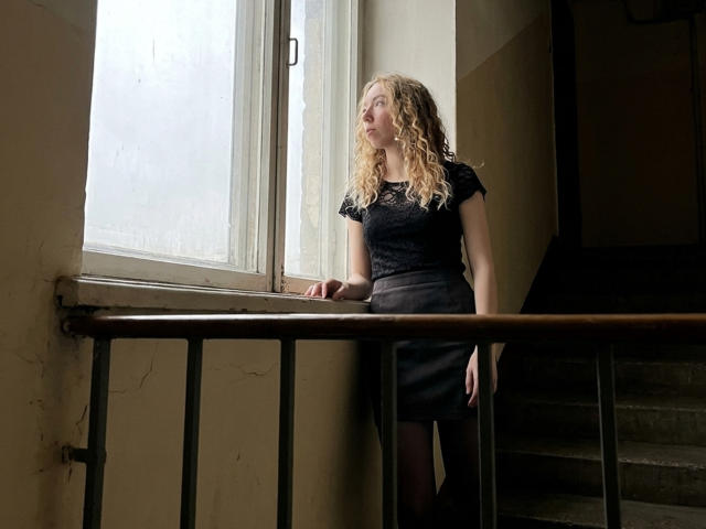 ClaireSpisak stands by a window, wearing a black lace top and gray skirt, looking outside with curly blonde hair, arms resting on a wooden railing.
