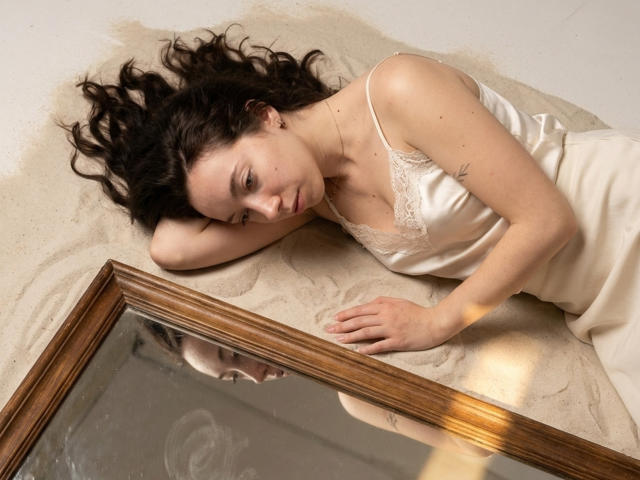 ClaireSpisak lies on sand, wearing a white lace-trimmed satin dress, with her dark curly hair spread out, hand on a mirror's wooden edge, reflecting her face.
