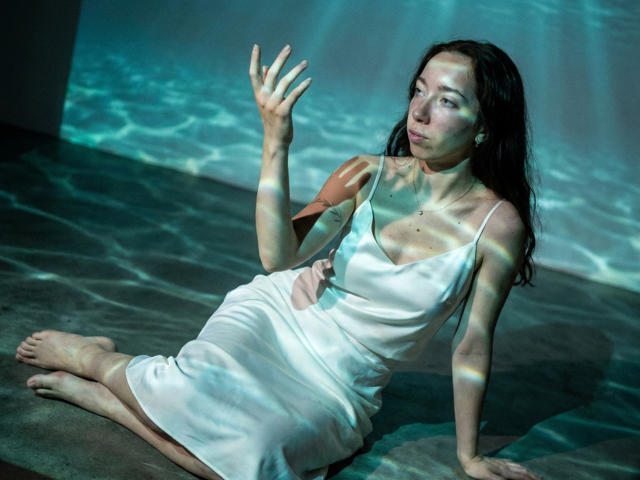 ClaireSpisak with wet dark hair, white silky dress, bare feet, sitting on floor, waving hand, sunlight and water reflections, small tits, cute, sexy, light skin, blue-green light.