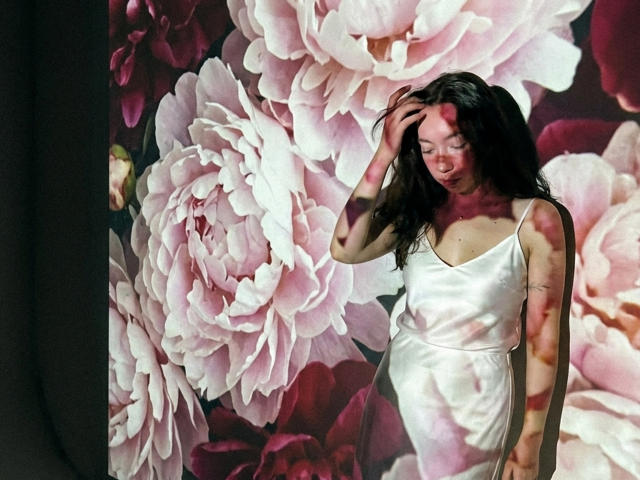 ClaireSpisak with long dark hair, wearing a white satin dress, standing in front of pink floral wallpaper, hand in hair, red paint splatters on her arm and chest.