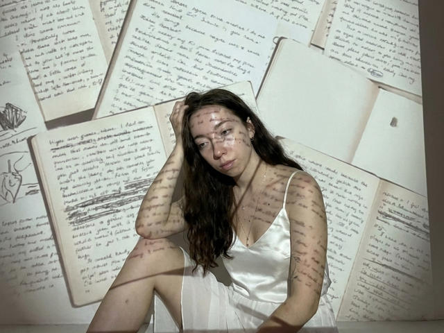 ClaireSpisak, sitting with a white satin dress, has long brown hair, and shadow patterns on her face and body. Her tits are small, and she has a pensive look.