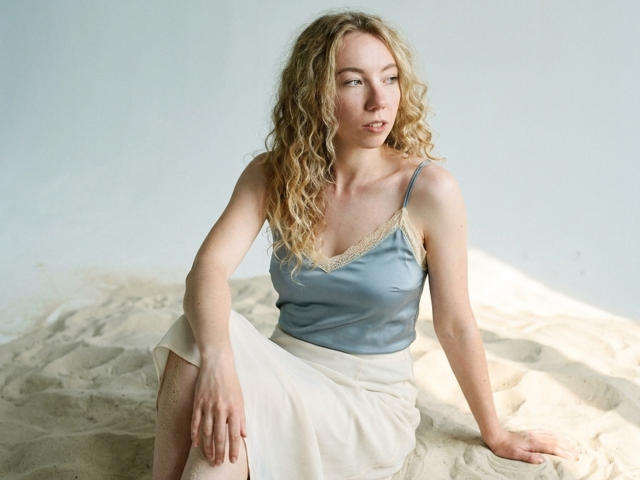 ClaireSpisak with curly blonde hair, blue satin top, and white skirt, sitting on sandy surface, looking to the side, soft natural light, small tits, pale skin, serious expression.
