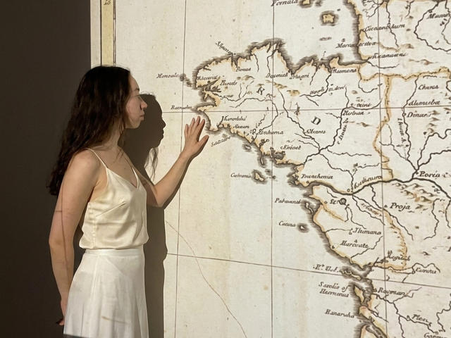 ClaireSpisak with long brown hair, in a silky white top and skirt, touches an old map of Europe, her hand near France. She stands in soft sunlight.