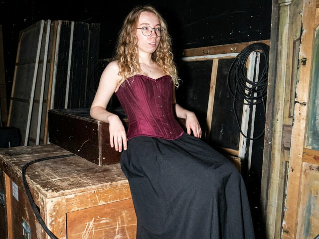 ClaireSpisak with curly blonde hair, glasses, and a maroon bustier, sitting on a wooden crate, wearing a black skirt, in a dimly lit, rustic room.