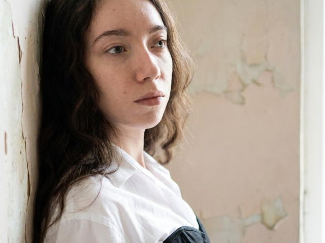 ClaireSpisak has wavy brown hair, light skin, and blue eyes. She's wearing a white shirt with a dark blue cross-strap, leaning against a worn, pale wall.