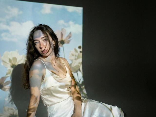 ClaireSpisak with long brown hair, wearing a white satin dress, sitting in shadow with light filtering through floral wallpaper, showing her tits and a subtle smile.
