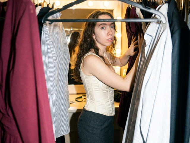 ClaireSpisak with wavy brown hair, wearing a cream corset and gray skirt, standing in a clothes rack, holding a white shirt, looking at the camera with a neutral expression.