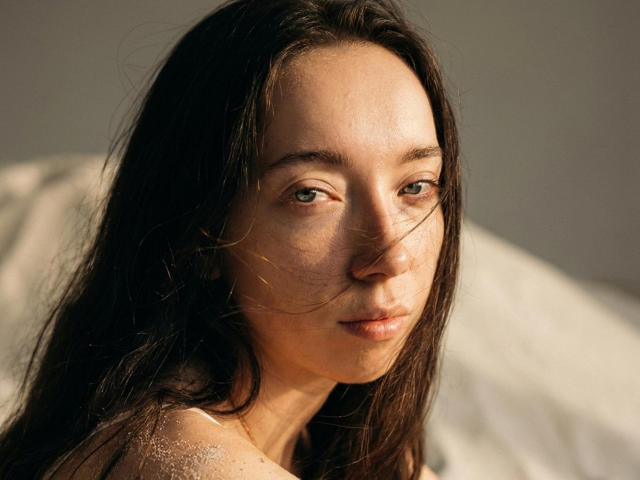 ClaireSpisak has long dark brown hair, green eyes, and a slightly dirty face with sand. She's looking at the camera with a calm expression, soft sunlight on her.