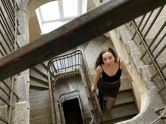 ClaireSpisak with tits in a black lace top and brown skirt, climbing a spiral stone staircase, looking up with a slightly open mouth, holding a railing.