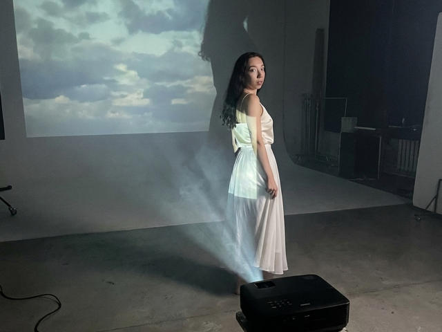 ClaireSpisak in a white, sleeveless, backless dress with a flared skirt, standing with her ass to the camera, illuminated by a projector casting a cloudy sky on the wall.