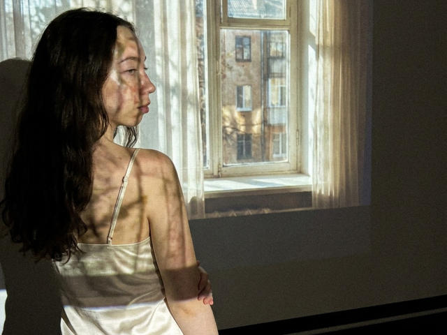 ClaireSpisak with long dark hair, white tank top, shadowed face, and arms, standing near sunlit window, looking outside at building. Her tits and arms cast in shadows.