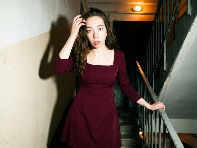 ClaireSpisak with wavy brown hair, wearing a red dress, standing in a dimly lit stairway, hand in hair, other hand on railing, looking up with a slight smile.