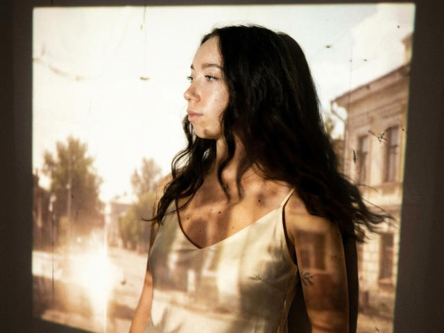 ClaireSpisak with long, wavy dark hair, wearing a gold tank top, has a pensive look, light skin, and a small tattoo on her right shoulder.
