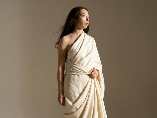 ClaireSpisak with long dark hair, wearing a white toga-like dress, holding the fabric, standing, looking to the side, neutral expression, soft lighting, beige background.