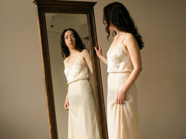 ClaireSpisak stands in front of a mirror, gazing at her reflection. She wears a silky, cream-colored dress, showing her tits and ass, with wavy dark hair.