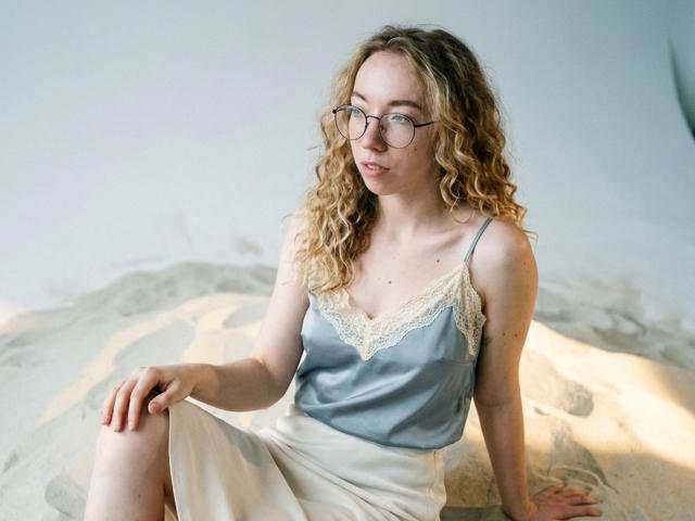 ClaireSpisak with curly blonde hair, wearing glasses, a blue satin top with lace, and a white skirt, sitting on sandy ground, looking slightly to the side.