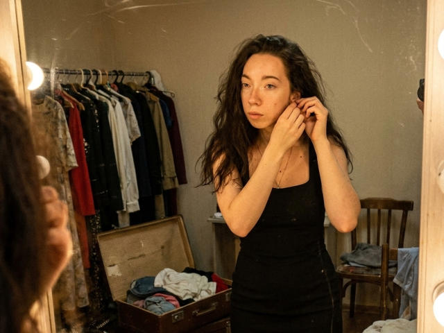 ClaireSpisak with messy dark hair, wearing a black tank top, stands in front of a mirror, hands near her face, looking slightly annoyed. Clothes hang on a rack behind.