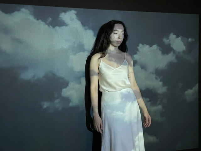 ClaireSpisak with long black hair, wearing a white satin dress, standing in shadow with cloudy light patterns on her skin and dress, looking down, hands by her sides.