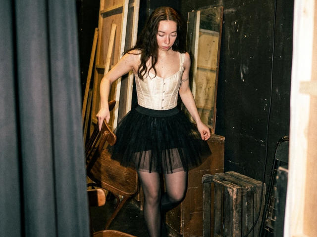 ClaireSpisak with long brown hair, white corset, black tutu, and sheer black stockings, holding wooden door, standing in dark, cluttered room, looking down, legs slightly apart.
