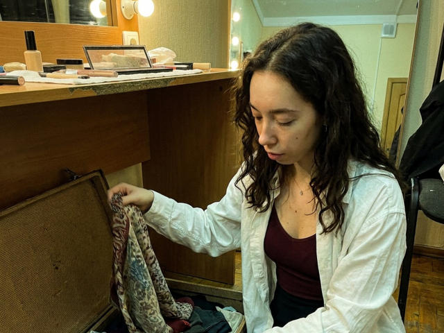 ClaireSpisak with wavy brown hair, white shirt, and maroon top, reaching into an open brown suitcase, holding patterned clothes. Her face is focused and slightly downcast.