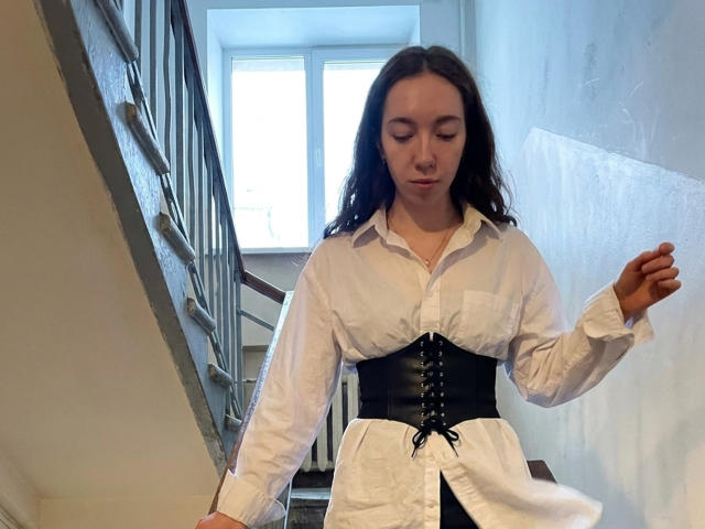 ClaireSpisak with wavy dark hair, wearing a white button-up shirt and black corset, standing on stairs, eyes closed, right hand raised, left hand on railing.