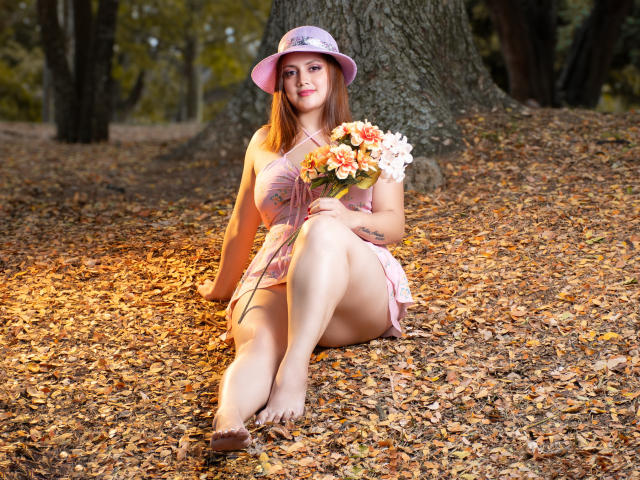 RoxanneBreats with pink hat, holding flower bouquet, sitting on autumn leaves, legs crossed, showing her ass, pink dress, bare feet, tattoo on left arm, smiling in forest.