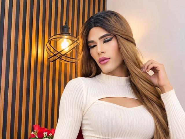 Scarleth Jojhansom, avec de longs cheveux bruns ondulés, un pull col roulé blanc texturé, un décolleté, du rouge à lèvres rose et un eye-liner noir, se tient à côté d'une ampoule suspendue..