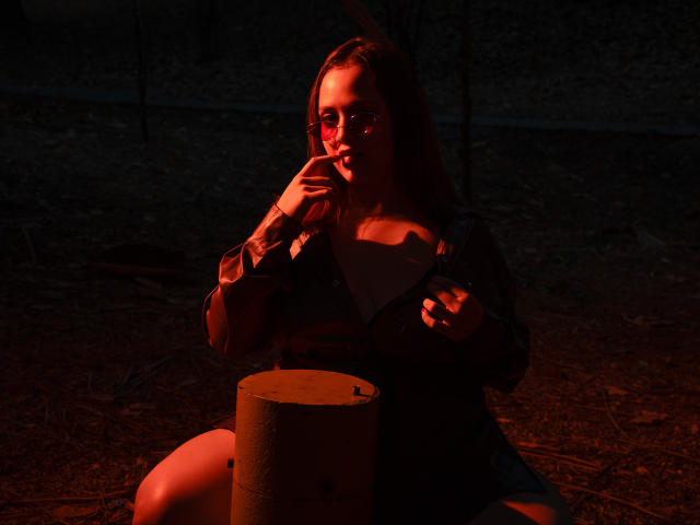 RoxanneBreats with brown hair, red eyes from dark light, finger to lips, dark jacket, white top, seated, hand on cylinder, dark forest background, red-orange light.