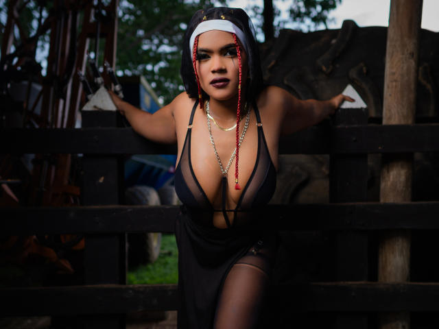 AliceJelanni with big tits, wearing a nun outfit, black sheer dress, red braids, chain necklace, leaning on a black wooden fence, looking at the camera, outdoors, dark makeup.