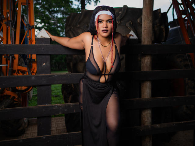 AliceJelanni leans on a black wooden fence, wearing a sheer black dress with deep cleavage, showing her large tits, and a nun's headpiece, with red beads in her hair.