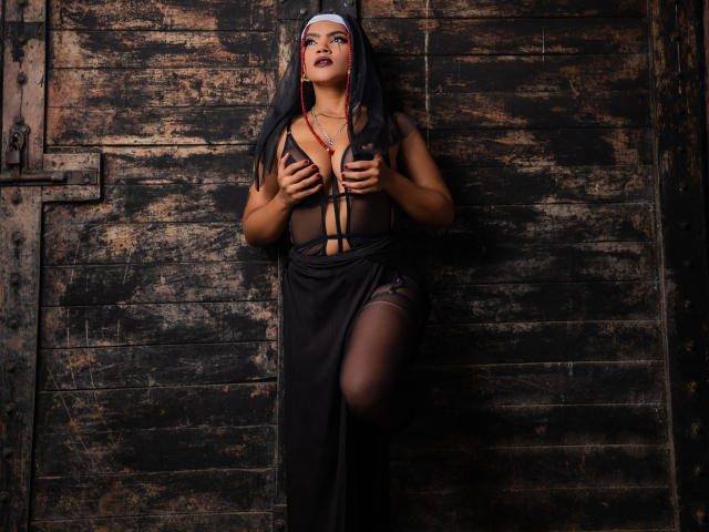AliceJelanni with dark skin, wearing a black sheer top, nun's veil, and red earrings, covering her big tits with her hands, against a worn wooden door.