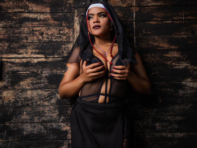 Alice Jelanni indossa un abito da suora nero con una parte superiore trasparente, coprendosi il seno con le mani, una collana di perline rosse, rossetto scuro e un velo nero, sullo sfondo di un muro di legno grezzo..