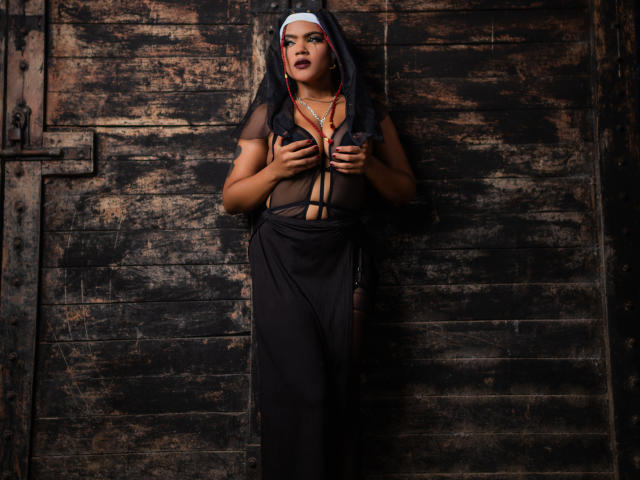 Alice Jelanni in un abito nero trasparente, che tiene i seni coperti da un velo da suora, indossa una collana, è appoggiata a un muro di legno sporco e appare seducente con il rossetto scuro..