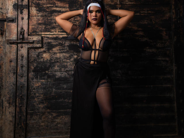 AliceJelanni with dark skin, wearing a nun's habit and sheer black lingerie, exposing her tits and pussy, standing against a rustic wooden door, hands behind her head.