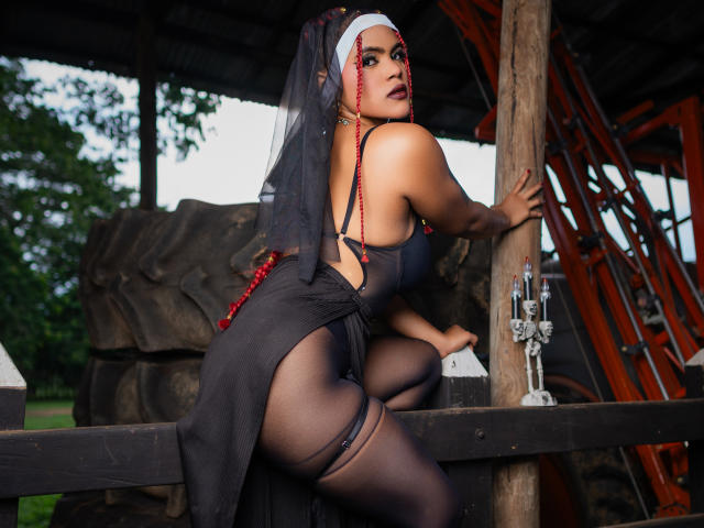 AliceJelanni in un abito nero da suora, calze velate, perline rosse, velo bianco, che guarda indietro, sedere in evidenza, appoggiata a un palo di legno, reggiseno nero, seni visibili, ambientazione esterna, candelabro a destra..
