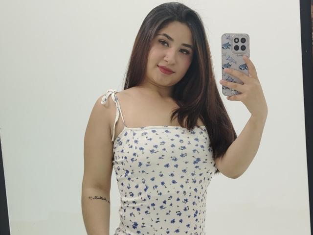 Julhiana mit langen, braunen Haaren, trägt ein weißes Kleid mit blauen Blumen und macht ein Selfie mit ihrem Handy. Ihre Brüste sind klein, und sie hat ein Tattoo am Arm..