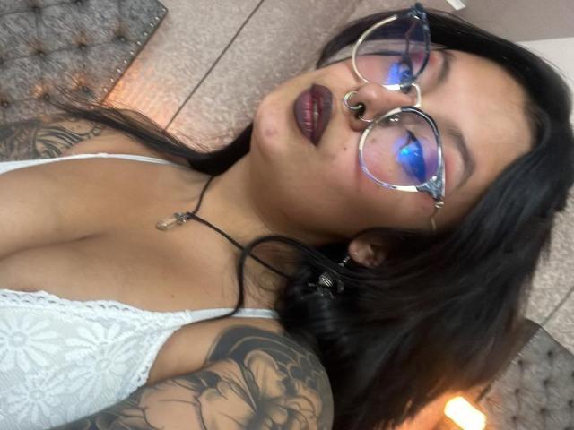 GabyJoy mit Tattoos, dunklem Lippenstift, Nasenring und blauem Lidschatten, trägt eine Brille und eine weiße Spitzenbluse und zeigt ihre Brüste. Ihr schwarzes Haar ist offen und sie schaut in die Kamera..