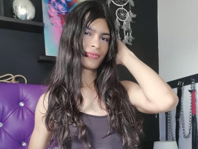 LunarisSmith has long, wavy brown hair, purple eyeshadow, and a pink lip ring. She's wearing a black tank top, with her right hand in her hair. Boobs visible.